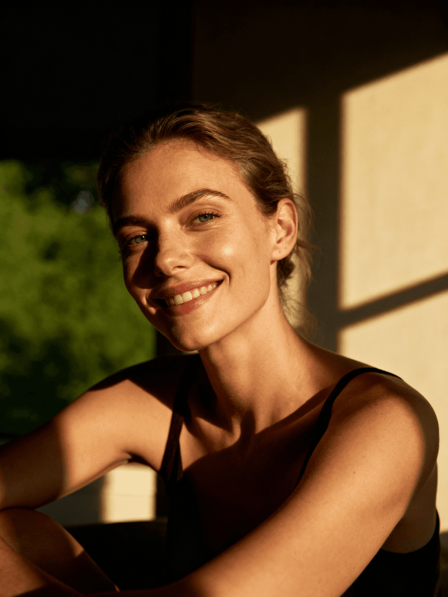 Woman smiling in warm sunlight with blurred green background.